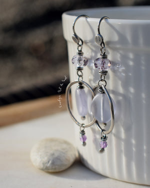 Handcrafted silver earrings with lilac and lavender beads, displayed on a white ceramic cup with a smooth stone in the background.