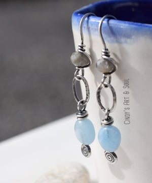 Silver earrings with pale blue aquamarine stones and gray labradorite stones. The earrings have silver hoops in the middle and spiral accents on the lower part. 