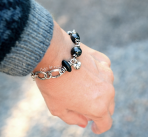 A close-up of a person's wrist wearing a silver chain bracelet with large black beads and a small silver star charm, set against a blurred outdoor background. The person is dressed in a gray, textured sleeve.