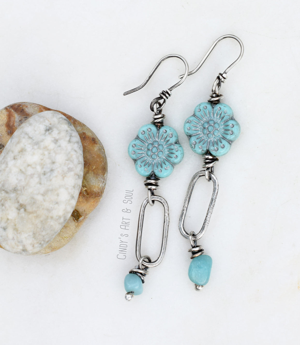 A pair of silver earrings featuring turquoise flower-shaped beads and silver oval links with small turquoise dangles at the bottom, displayed on a white background next to a smooth stone.