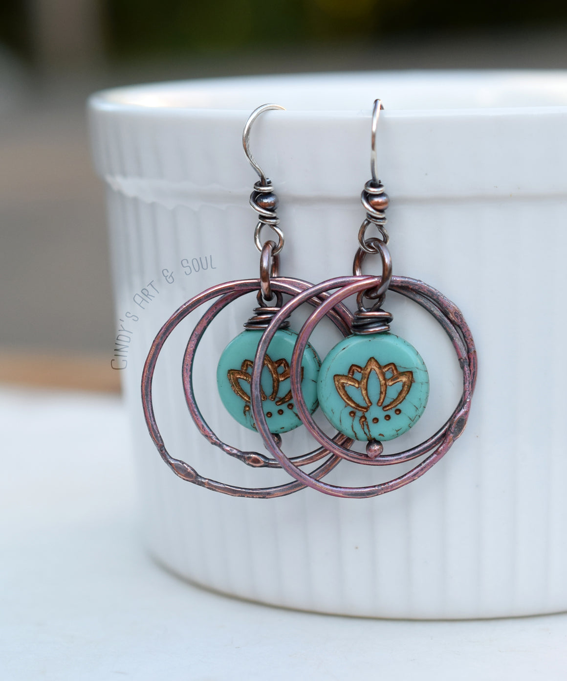 Copper hoop earrings with turquoise and bronze lotus beads, hanging from a white cup. 