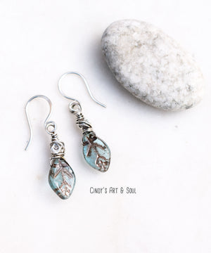 Blue leaf earrings with silver beads on a white background with a gray marbled stone sitting on the upper right side of the image.