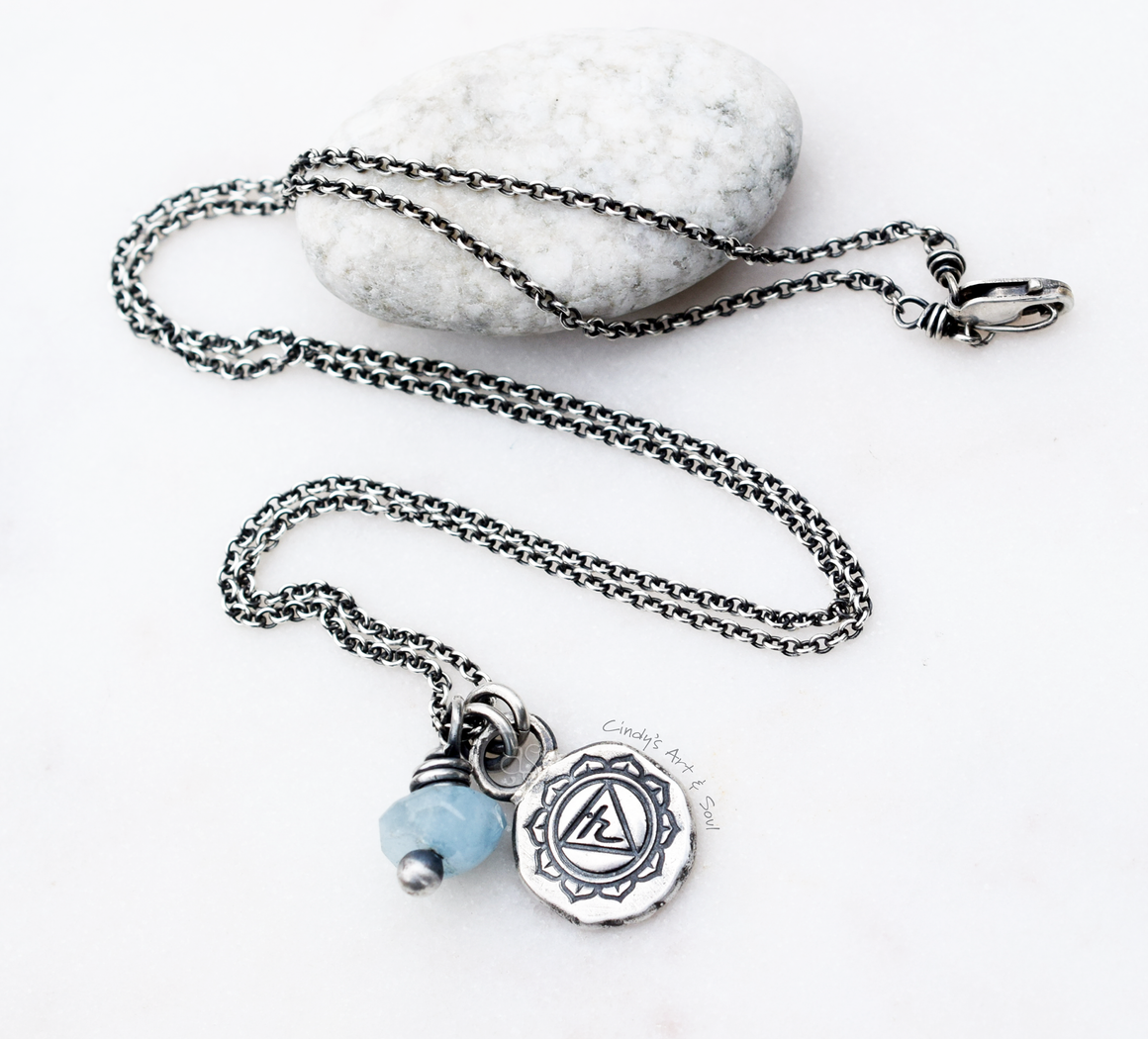 
A silver chain necklace with a small blue bead and a round silver pendant featuring an engraved symbol, displayed on a white background with a smooth white stone.