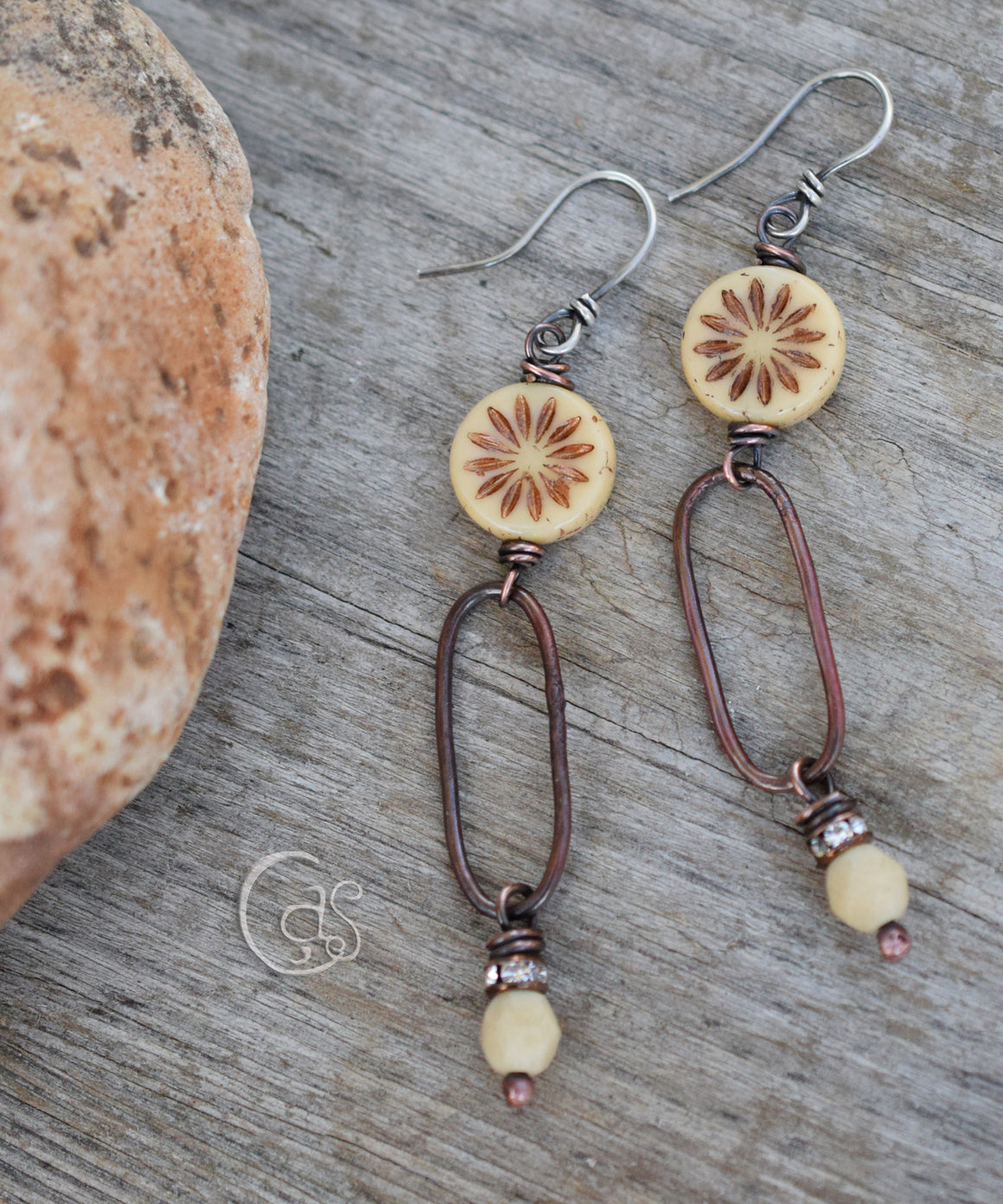 Copper and ecru flower beads with copper hoops and rhinestone dangles with silver ear wires on a wood background, next to an orange rock. 