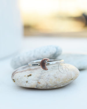 A silver ring with a copper crescent moon sitting on a beige rock. The background is white.