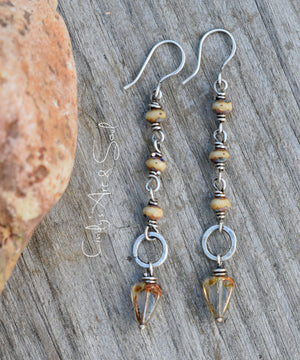 Silver earrings with small hoop rings and beads that are sand and crystal colored on a wood background next to an orange stone. 