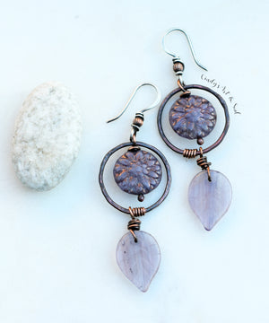 Alt text: "Handcrafted earrings featuring purple Czech glass flower beads, with circular copper hoops and wire wrapping, accented by light lavender glass leaf drops. The earrings are displayed on a light background next to a small white stone."