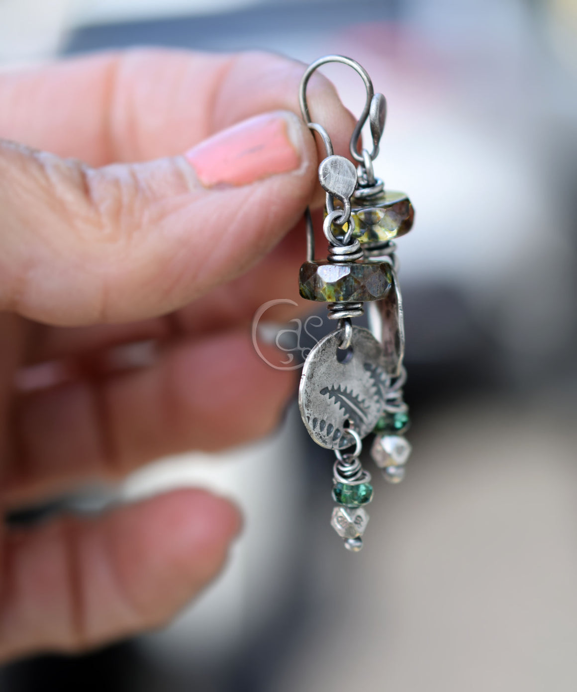 A hand holding green and silver earrings with a blurry background. 