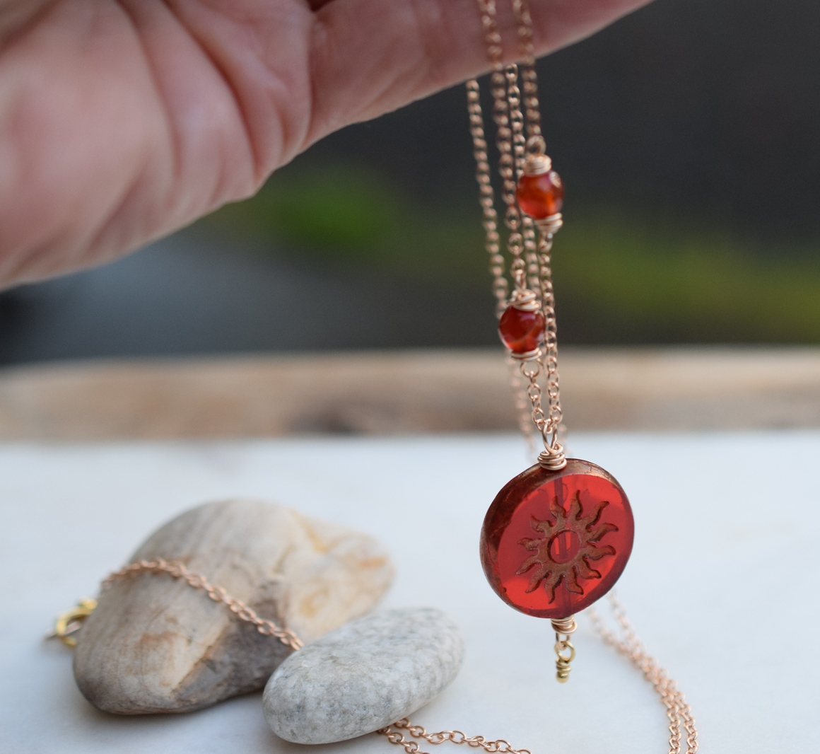 Hand holding a gold necklace with red beads and a sun pendant with a white background and small rocks at the bottom