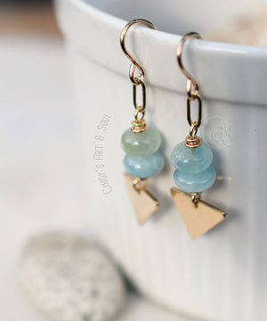 Gold Earrings with blue green stones hanging from a white cup and a small river stone sitting below.