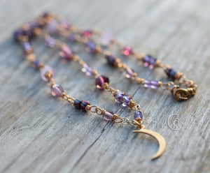 Gold necklace with purple beads and a gold crescent moon charm on a wood background. 