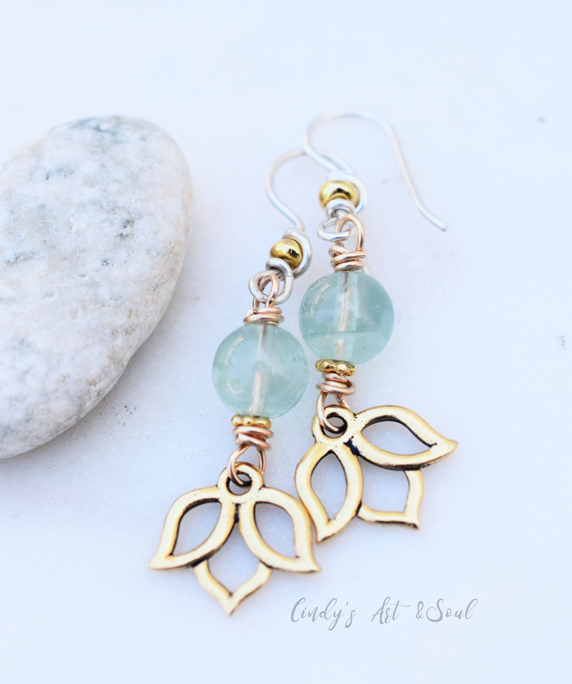 A pair of earrings featuring light green round beads, gold accents, and gold lotus flower charms at the bottom, displayed on a white surface next to a smooth white stone.