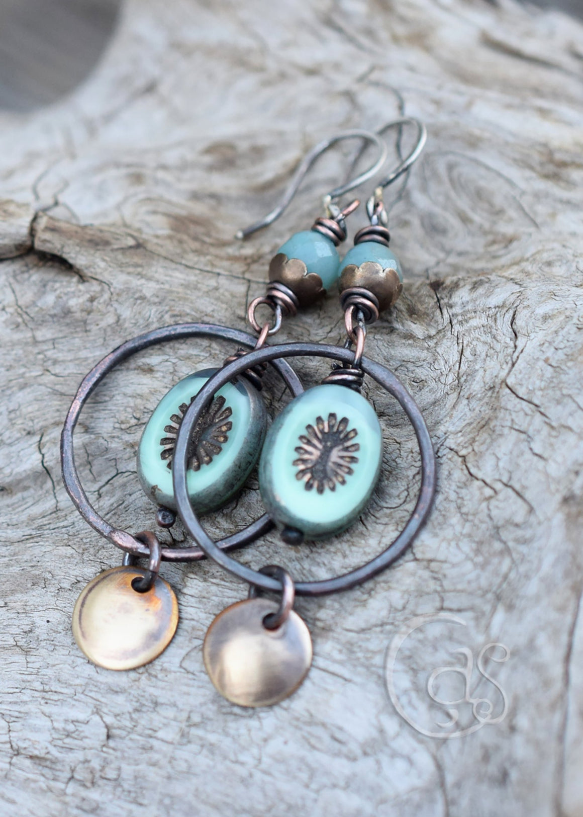 Rusty copper hoop earrings with blue beads that have golden star bursts on them. golden charms dangle below. The top of the earrings have blue stone beads with brass flower caps. The ear wires are silver. These earrings are laying flat on a piece of driftwood. 