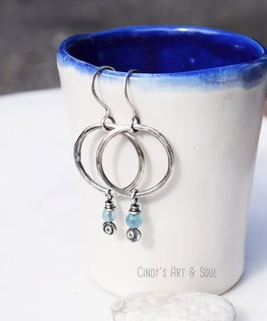 A pair of silver hoop earrings with small blue beads and decorative silver accents, hanging from ear hooks, displayed on the edge of a white ceramic cup with a blue interior. A small stone is placed at the base of the cup.