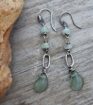 Pale green beaded silver earrings with darker green dangles below on a wood background next to a orange stone. 