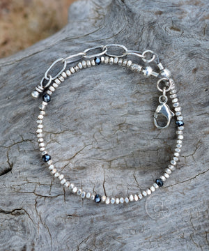 Bracelet with tiny silver beads and blue-black stones, with a chain end and lobster clasp, sitting on a piece of wood. 