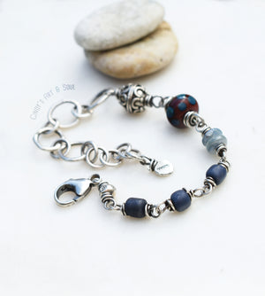 Handcrafted bracelet with a mix of blue and silver beads, connected by intricate silver wire links and featuring a lobster clasp closure. The bracelet includes textured beads, a large dotted focal bead, and delicate chain links, set against a minimalist background with smooth stones.