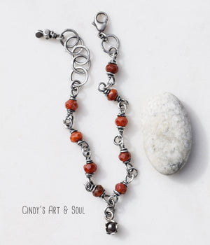 A beaded bracelet with orange mexican fire opal gemstones that has a silver chain. There is a flower charm at the center and a length of silver chain at the end. There is a gray and white stone next to the bracelet and the photo has a white background. 