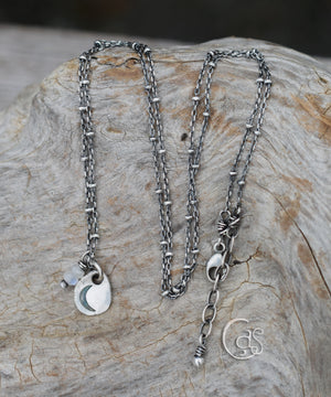 A silver necklace with a crescent moon charm and a moonstone charm on a beaded chain sitting on a wood background. 