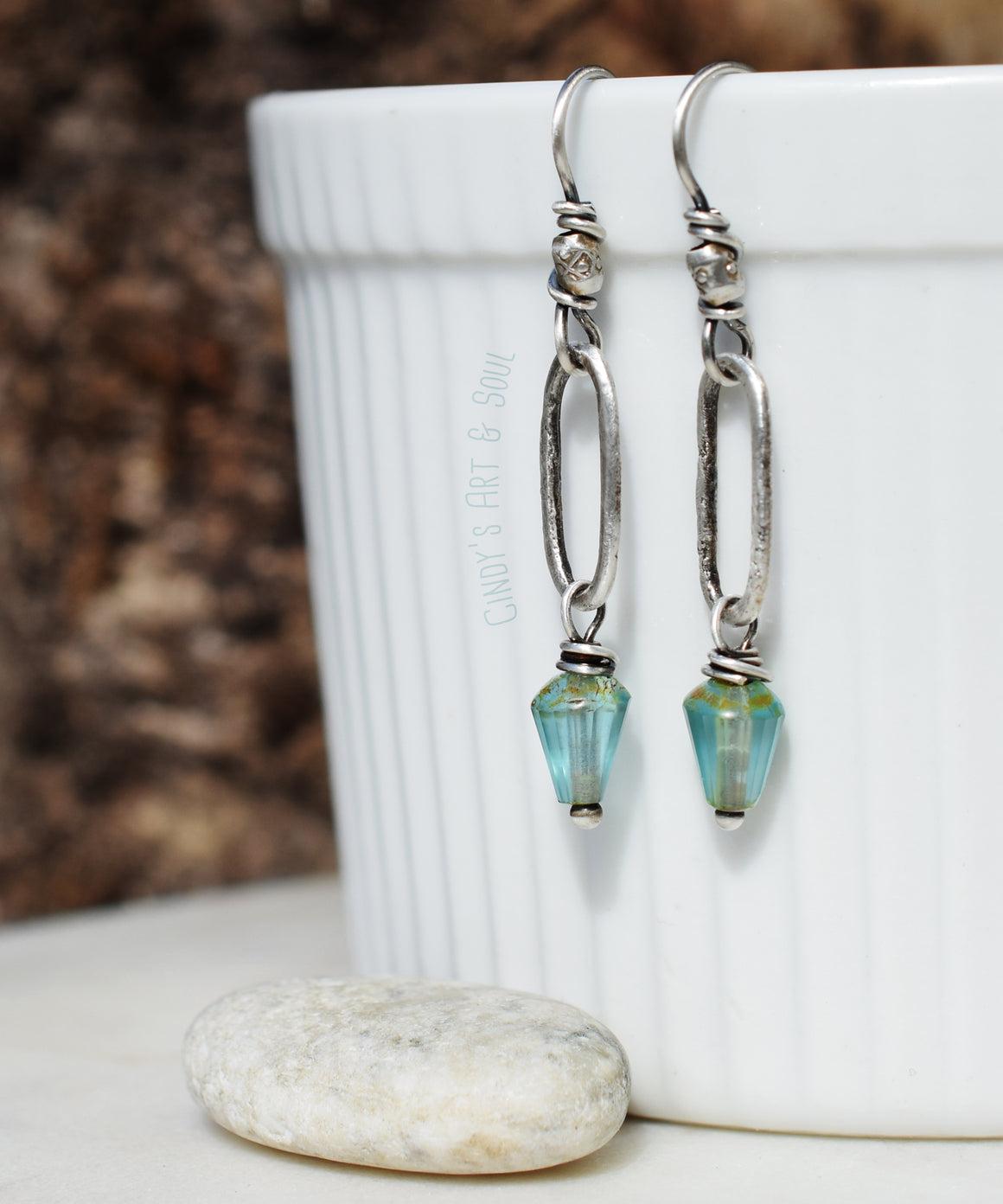 Silver hoop earrings are hanging from a white cup. The earrings have ocean-blue glass bead dangles. There is a white stone under them. The background is brown. 