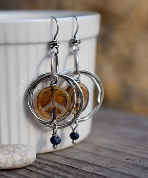 Handcrafted double silver hoop earrings with peace sign beads at the center, dangling from a white bowl sitting on a piece of wood with a small white stone. Blue beads dangle off the bottom of the hoops. 