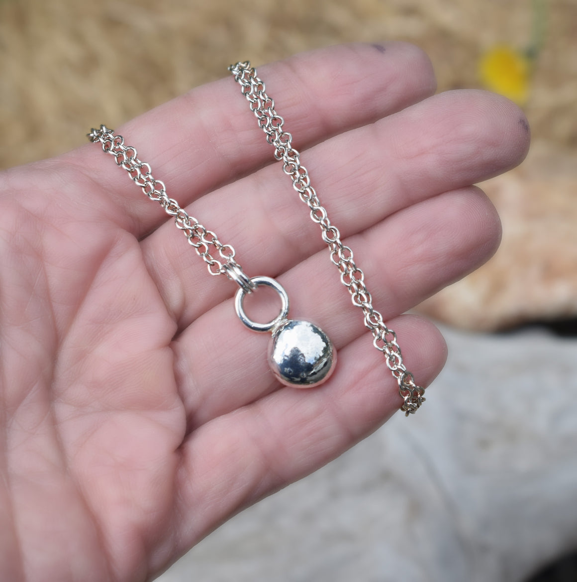 a hand showing the palm holding a Silver ball charm on a necklace chain.