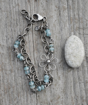 A silver and blue beaded bracelet with a flower sitting next to a stone on a wood background. 