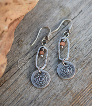 Silver earrings with peach stone dangles and solar plexus chakra symbol charms below oval hoops. They are on a wood background and there is a rock off to the left side. 