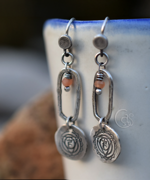 Silver Earrings with Solar Plexus Chakra Charms and small peach moonstones dangle in the upper center of silver hoops. These earrings are hanging from a white cup with a blue rim next to a blurry rock. 