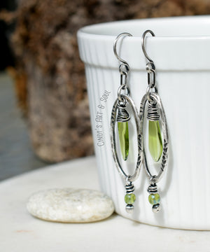 A pair of silver earrings with green glass teardrop beads encased in hammered oval silver frames, accented by small green beads at the bottom. The earrings are displayed hanging from a white cup, with a smooth stone placed nearby.