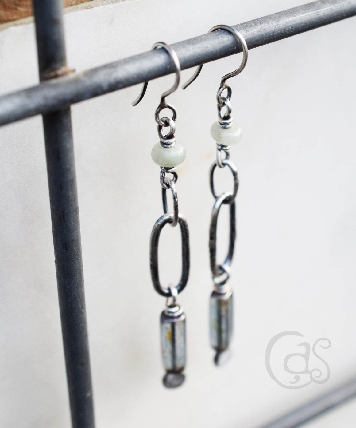 silver earrings with pale green stones and silver hoops and blue glass beads hanging from a metal wire on a white background