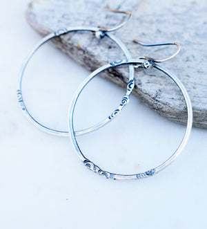 Silver hoop earrings with spiral designs on a white background next to a rock. 